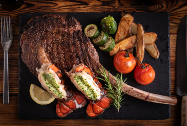 An aerial view of a grilled tomahawk steak and twin lobster tails served with potatoes and vegetables.