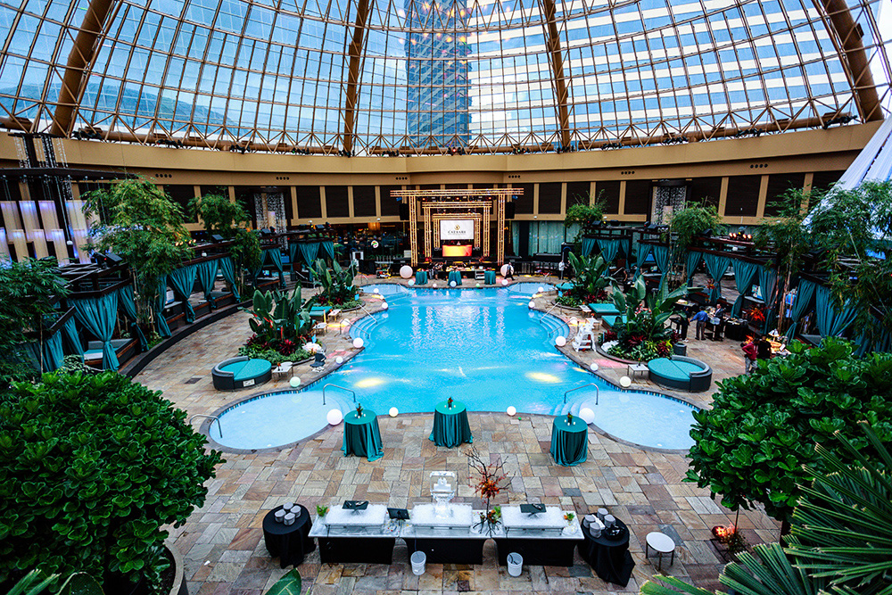 Aerial shot of indoor pool with plants and seating around it and a clear dome ceiling above.