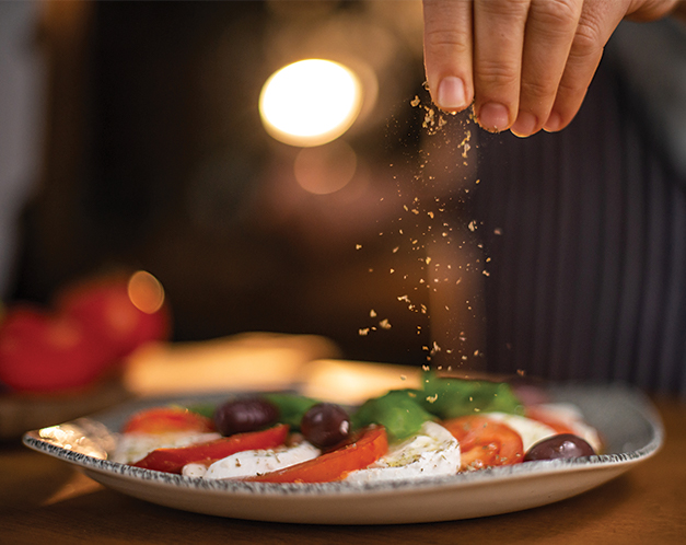 a person's hand sprinkling seasoning overtop tomato slices, mozzarella, and olives