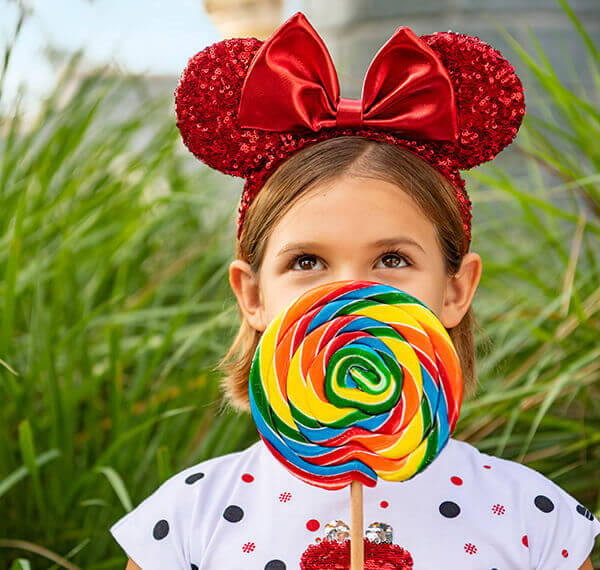 girl with bright red minnie mouse ears holding a colorful lollipop in front of her mouth