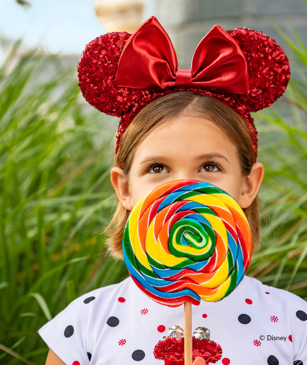 little girl wearing sparkly red Minnie Mouse ears and holding a large rainbow lollipop