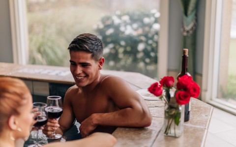 man and woman clinking their wine glasses while sitting in the hot tub with a vase of red roses on the side