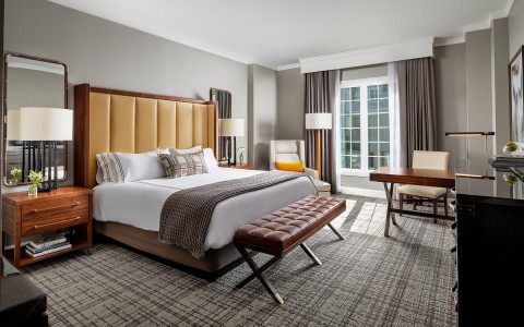 bed in room with a bench at the foot of it and french door style windows on the wall towards the right of the bed behind the desk and chair