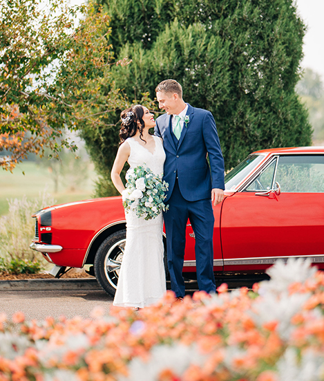 A married couple looking at each other in front of a classic red car