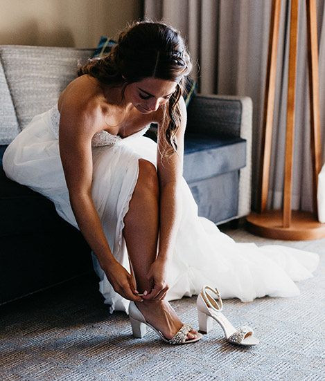 Bride sitting on a grey couch buckling her silver high heel shoe on