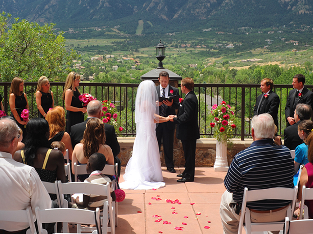 bride and groom getting married on a terrance outlooking a lush green valley in front of 2 rows of guest sitting in white chair and in the middle is red rose petals scattered on the ground