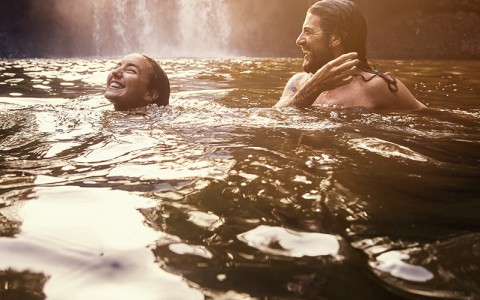 man and woman swimming playfully rising out of water at bottom of waterfall