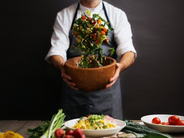 chef with blue apron holding a bowl where he is tossing salad