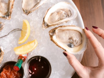 Hand holding an oyster above a dish of chilled oysters and sliced lemons with a tiny bottle of hot sauce