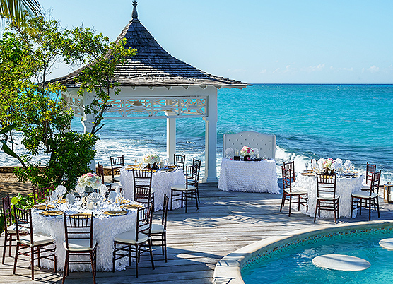round tables with white table cloths on a terrace with a gazebo and pool overlooking the ocean