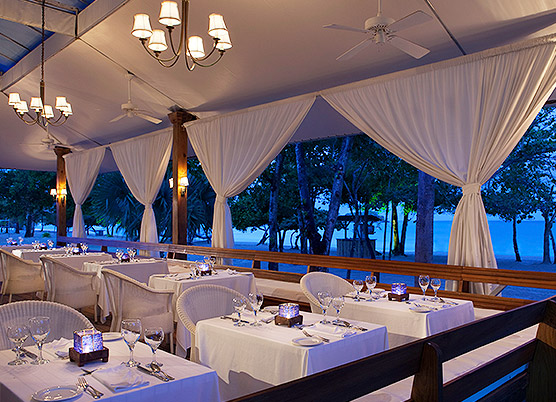 elegant covered outdoor dining area with white drapes, white tablecloths, and a view of the beach and ocean 