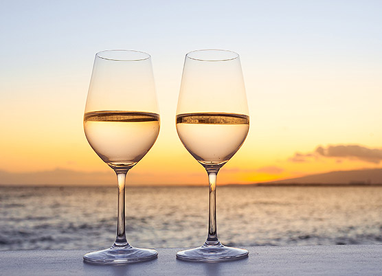 two glasses of white wine on a ledge in front of the ocean at sunset
