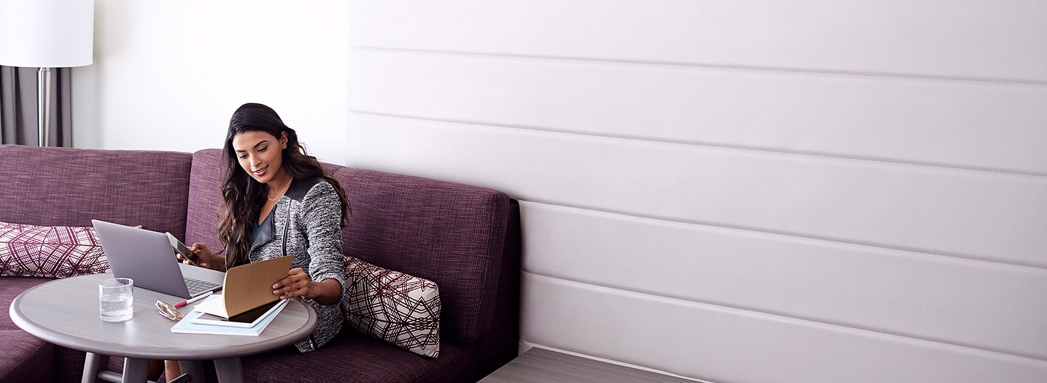 a woman working at a small table seated on a purple couch in the corner of a guest room
