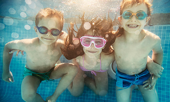 three kids under the water in a pool smiling with goggles on