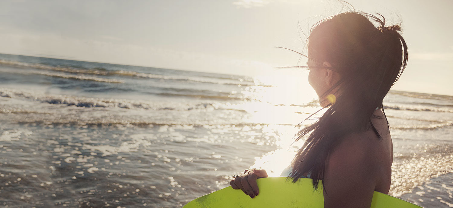 a woman holding a neon skimboard walking towards the ocean water with the sun gleaming behind her