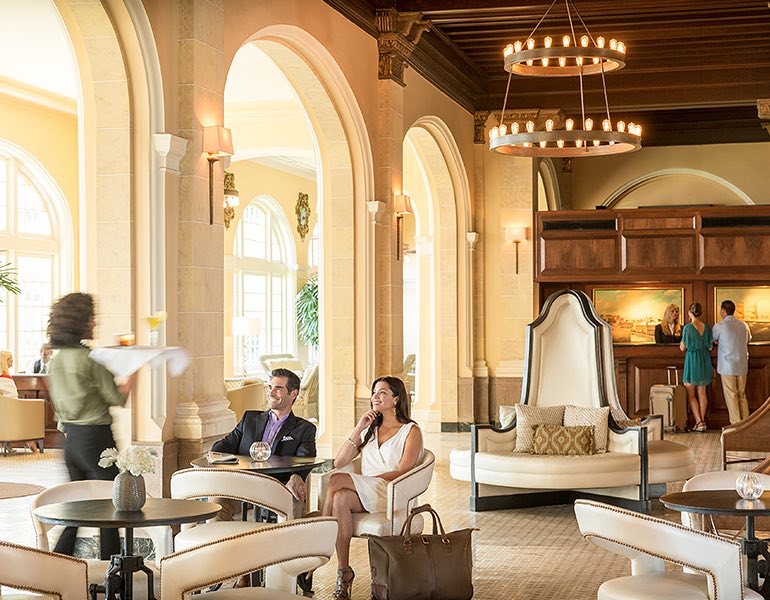 A waitress walks towards a couple seated in the lounge area carrying a tray of drinks