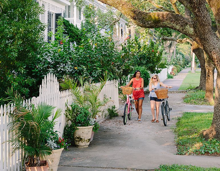Two women walk alongside their bicycles on a sidewalk next surrounded by greenery and white picket fences