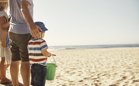 family on beach with dad holding his hand behind the little boy's hand while boy holds green bucket