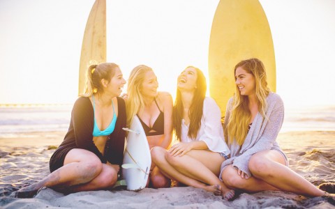 four young women sitting on beach laughing with surfboards behind them