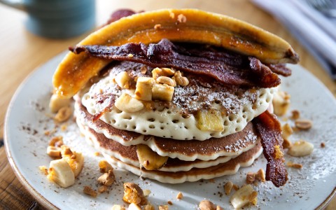 a stack of pancakes topped with sliced banana, nuts and bacon