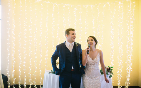 bride and groom giving a speech in front of a wall of twinkle lights