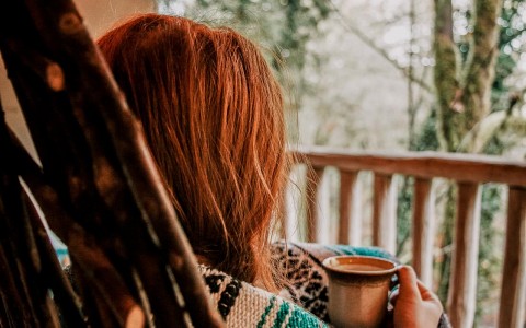 woman snuggled up in a blanket sitting outside on cabin patio drinking coffee looking into the woods