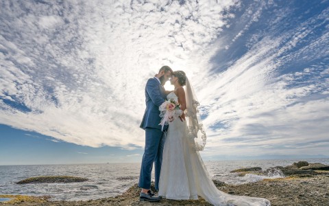 Panoramic view of a bride and groom hugging at the beach while the bride is holding a boutique of roses and a clear sky