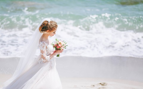 Bride walking at the beach with closed eyes while she is holding a boutique roses