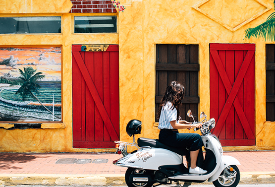 woman on a moped in front of a yellow building with red doors