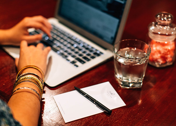 a woman typing on a laptop with a glass of water and pen and paper next to her