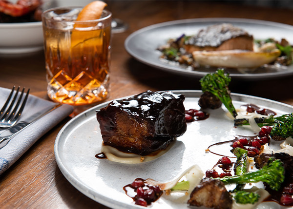 elegant steak dish with vegetables served on a white plate next to another elegant dish and a beverage