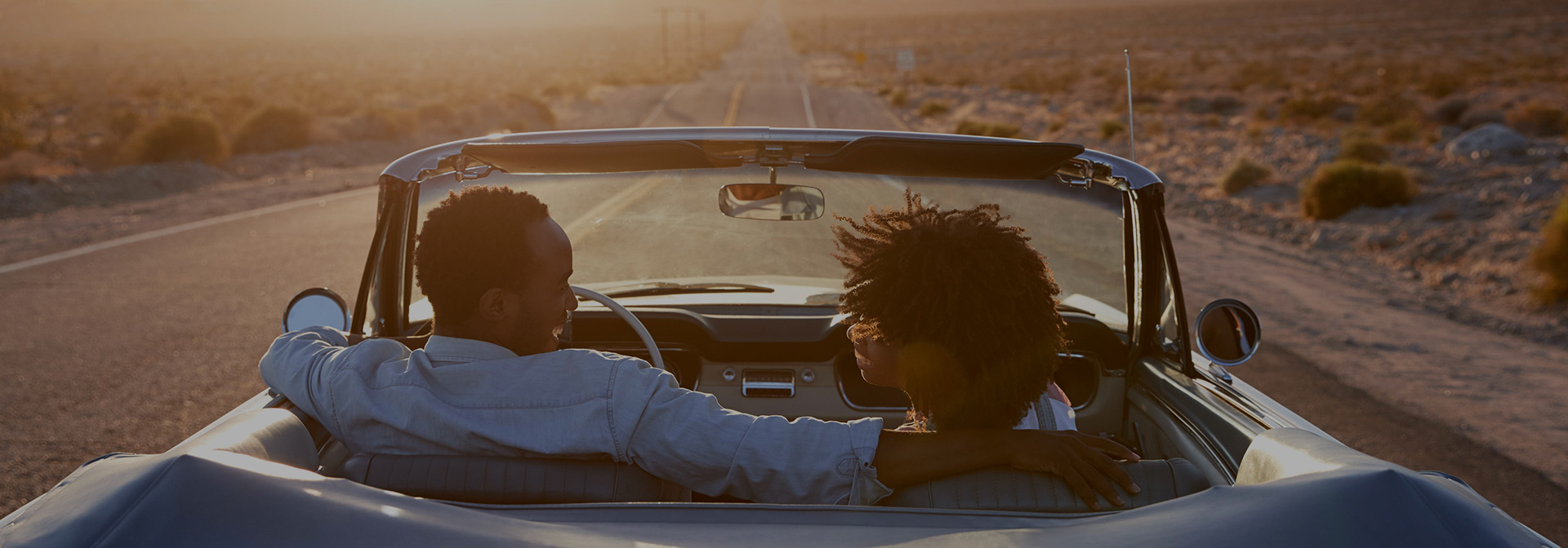 a man and woman riding in a convertible car while looking at each other at sunset