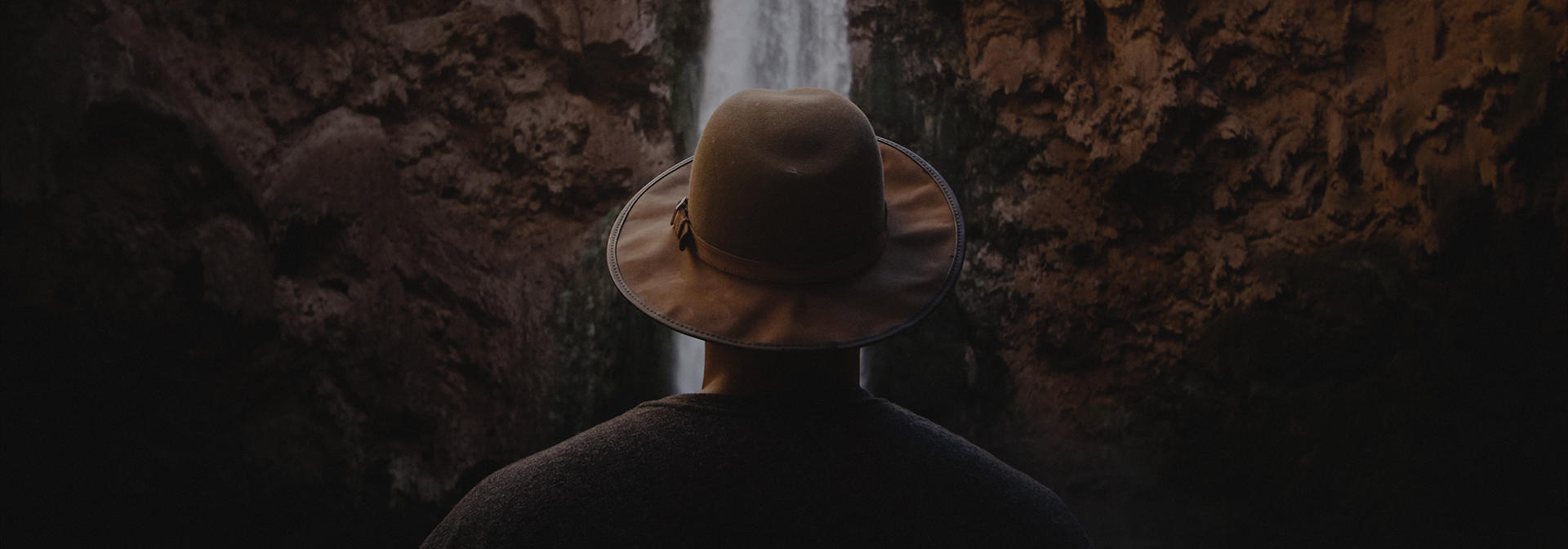 man wearing a fedora looks on a waterfall