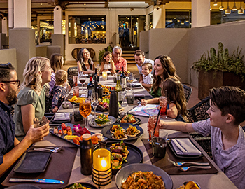 a large group of adults and kids dining outside on a terrace with the sun setting