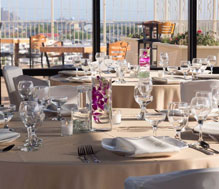 Event space housing several round tables with off white table clothes, covered chairs and formal dinner ware. Large glass wall opens to outdoor patio
