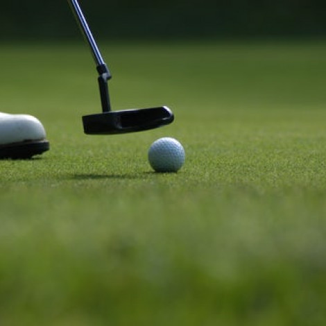 Golf putter hitting a golf ball on the green with white shoe also in shot