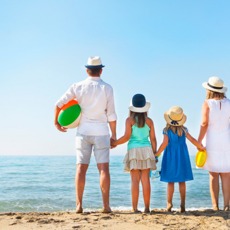 Family of four at the beach facing the water on the shore, they are carrying inflatable water toys