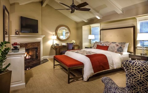 a bedroom with a brick fireplace and a bed with white bedsheets and a red and paisley printed throw and accent pillows
