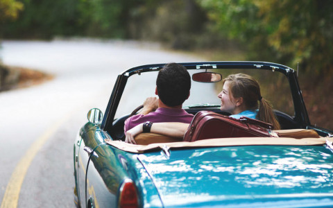 Couple in light blue convertible driving through street surrounded by greenery