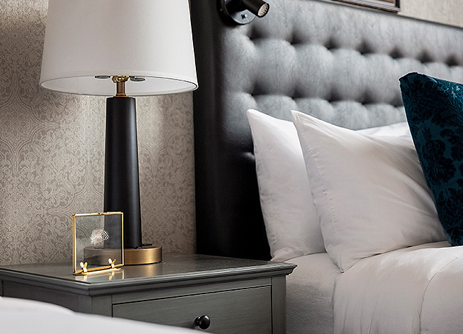 close up of a grey night stand ith lamp and bed with white sheets and black headboard