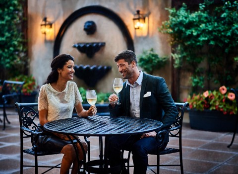 Man and woman drinking white wine on the patio with a water fountain and garden behind them 