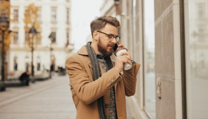 guy wearing winter outfit and glasses is taking a phone call while he is holding a large cup of coffee in the street