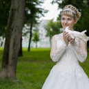 Blonde bride in white wedding dress holding a white dove on green grass next to trees