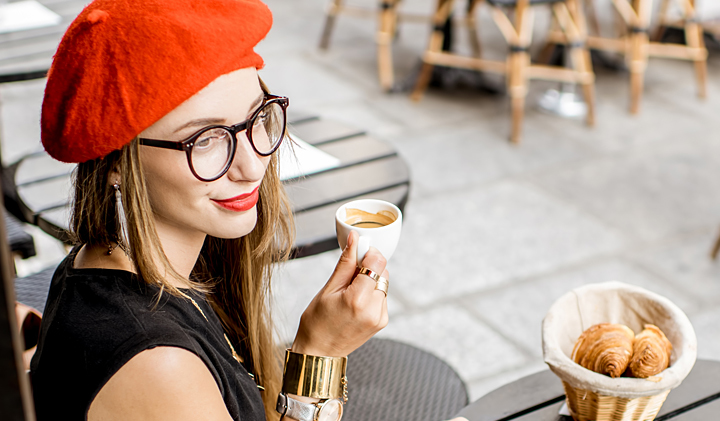 young woman wearing a red had and glasses sitting at a table drinking coffee