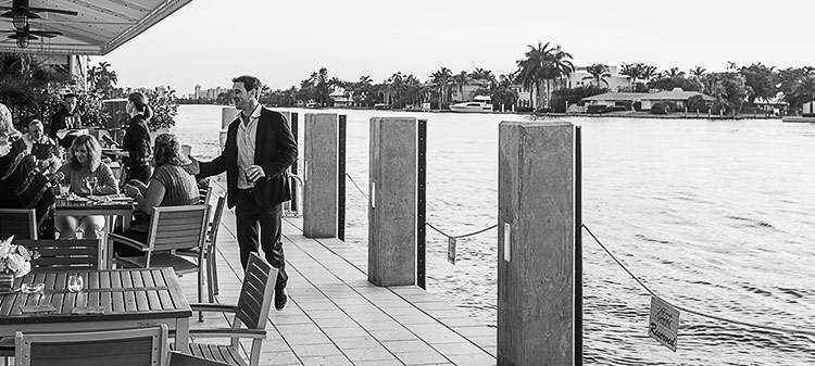 black and white image of a man walking towards a dining table on a covered patio area next to the water