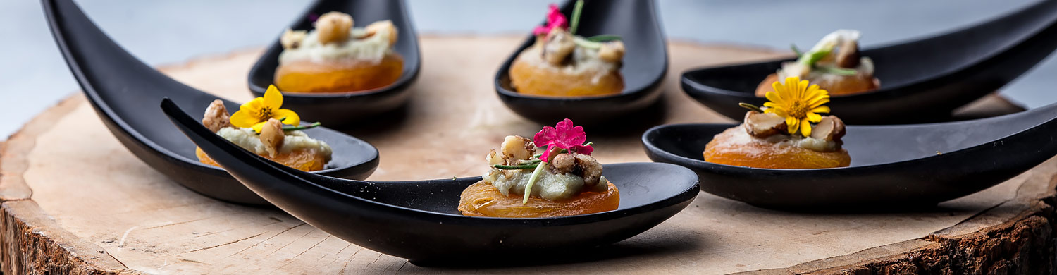 cheese crackers garnished with a small flower on individual black teardrop spoons on a rustic wooden slab