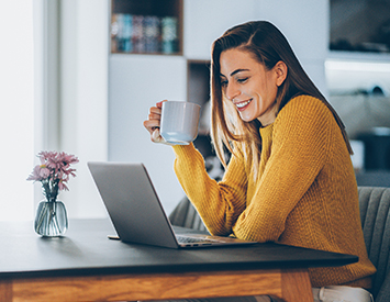woman in a yellow sweater drinking coffee and sitting with her laptop in front of her