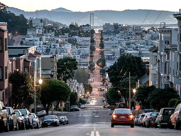 top of street hill looking down over a san francisco road with bridge in distance and cars lined on street