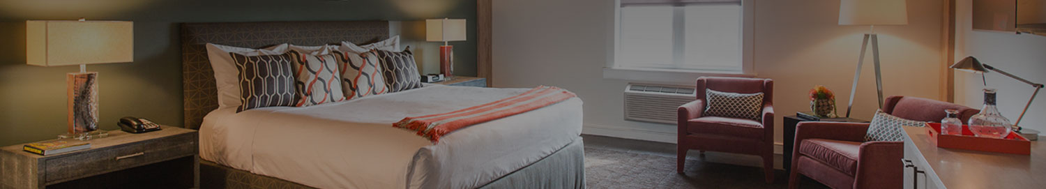 Interior of a king bed room with decorative pattern pillows, an orange throw on the bed, two nightstands, dresser, and a seating area with two chairs