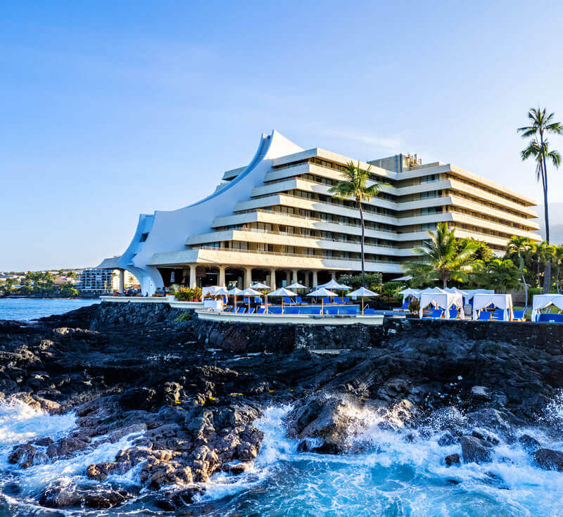 blue waves crashing upon volcanic rock next to Hawaiian hotel resort with oceanside pool